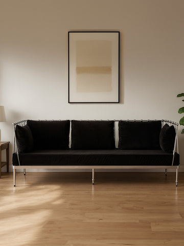 Black velvet three-seater sofa with a sleek silver frame, positioned against a light-colored wall. The sofa features three plush cushions and is complemented by a minimalist artwork hanging above it. A side table with a lamp and a potted plant are visible in the background, enhancing the modern living space ambiance