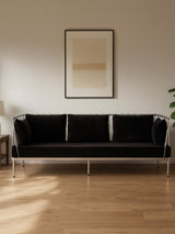 Black velvet three-seater sofa with a sleek silver frame, positioned against a light-colored wall. The sofa features three plush cushions and is complemented by a minimalist artwork hanging above it. A side table with a lamp and a potted plant are visible in the background, enhancing the modern living space ambiance