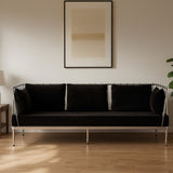 A sleek three-seater sofa upholstered in black velvet, featuring a modern silver frame. The sofa has three plush cushions and is positioned against a light-colored wall, with a minimalist artwork above it. A floor lamp and a potted plant are visible in the surrounding space, enhancing the contemporary aesthetic