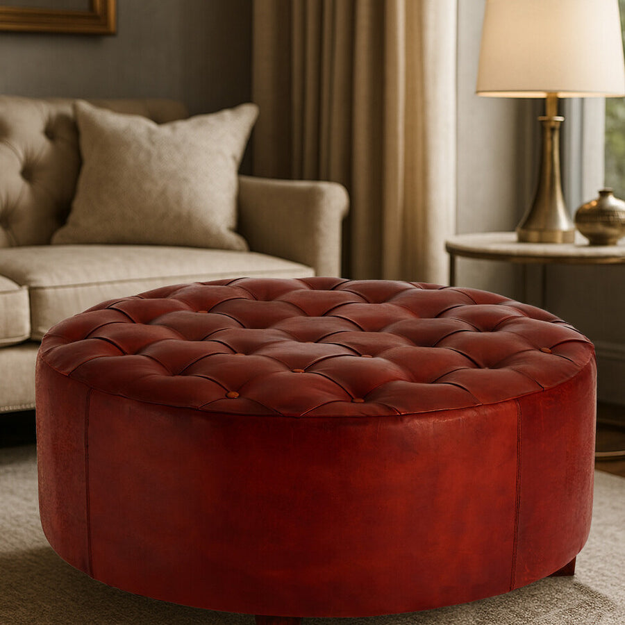 Round button tufted stool in antique brown leather, featuring a plush top with deep button detailing and a smooth, rounded base. The stool is positioned in a cozy living room setting, with a soft rug beneath it and a warm lamp in the background, creating an inviting atmosphere