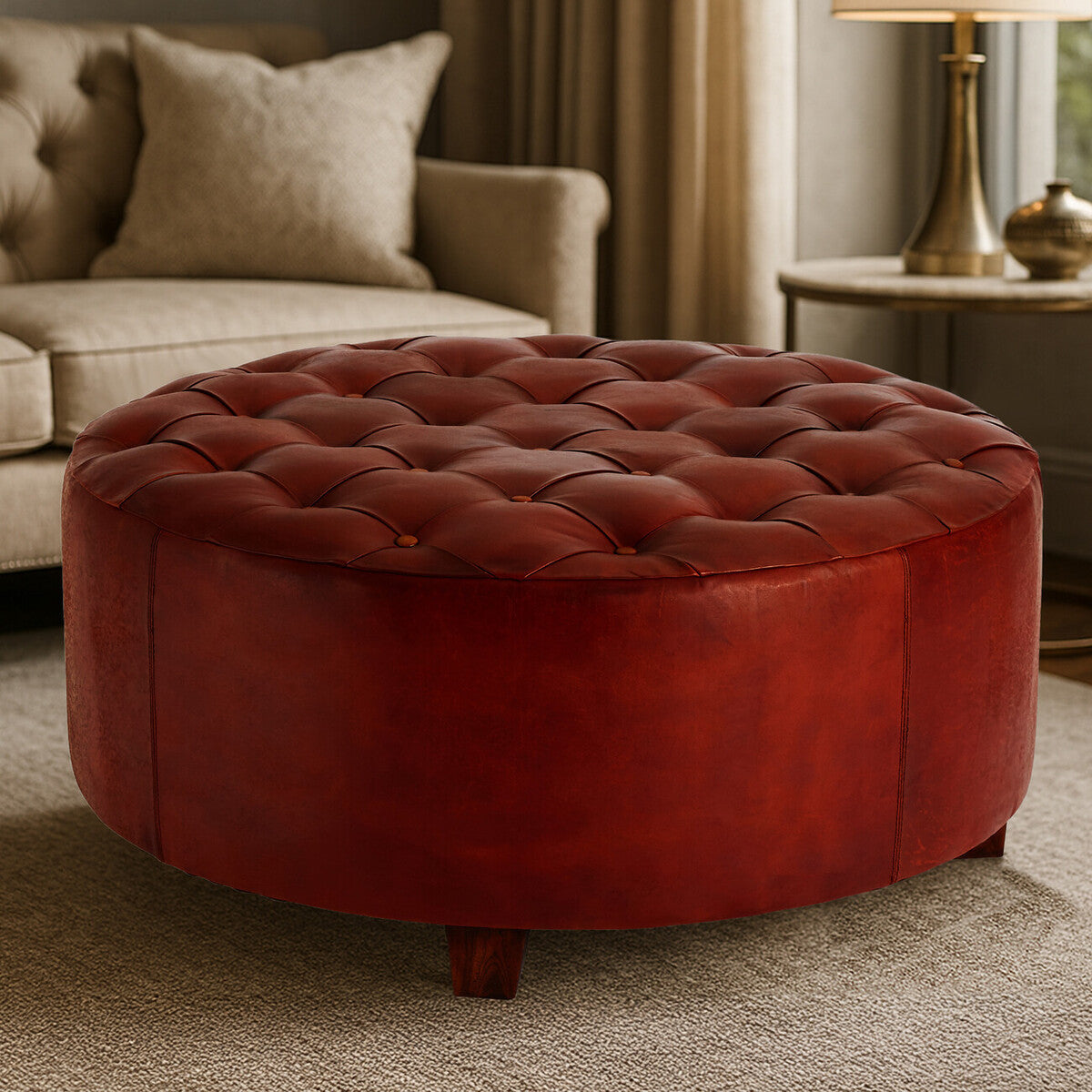 Round, button-tufted stool in antique brown leather with a smooth finish and rounded edges, featuring a plush top with evenly spaced buttons. The stool is positioned on a light-colored carpet, with a soft sofa and a lamp visible in the background, creating a cozy living room setting