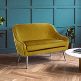 Mustard yellow velvet two-seater sofa with a curved backrest and sleek metal legs, positioned in a room with teal walls and wooden flooring, accompanied by a round coffee table and potted plants