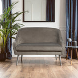 Grey velvet two-seater sofa with a sleek design, featuring rounded edges and a comfortable seat. The sofa is positioned in a bright room with wooden flooring, accompanied by a small side table and a decorative plant. A large mirror hangs on the wall behind the sofa, and blue curtains frame a window, adding a touch of elegance to the setting