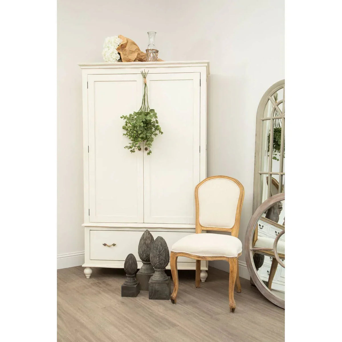 Cream wooden wardrobe with drawer beside cream upholstered dining chair and decorative mirror