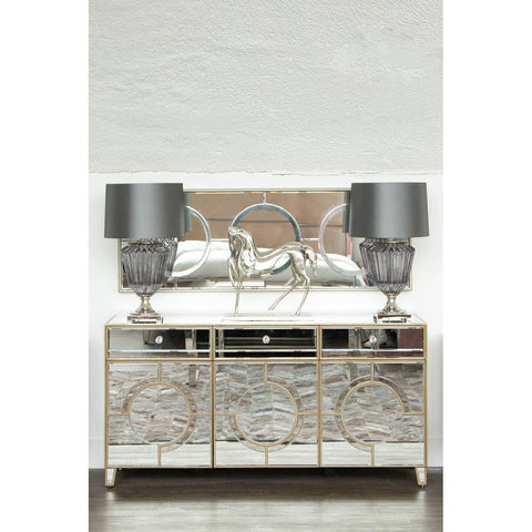 Champagne gold and mirrored sideboard featuring intricate geometric designs, two drawers, and two cabinet doors, topped with two elegant gray lamps. The background is a light-colored wall, enhancing the sideboard's reflective surfaces