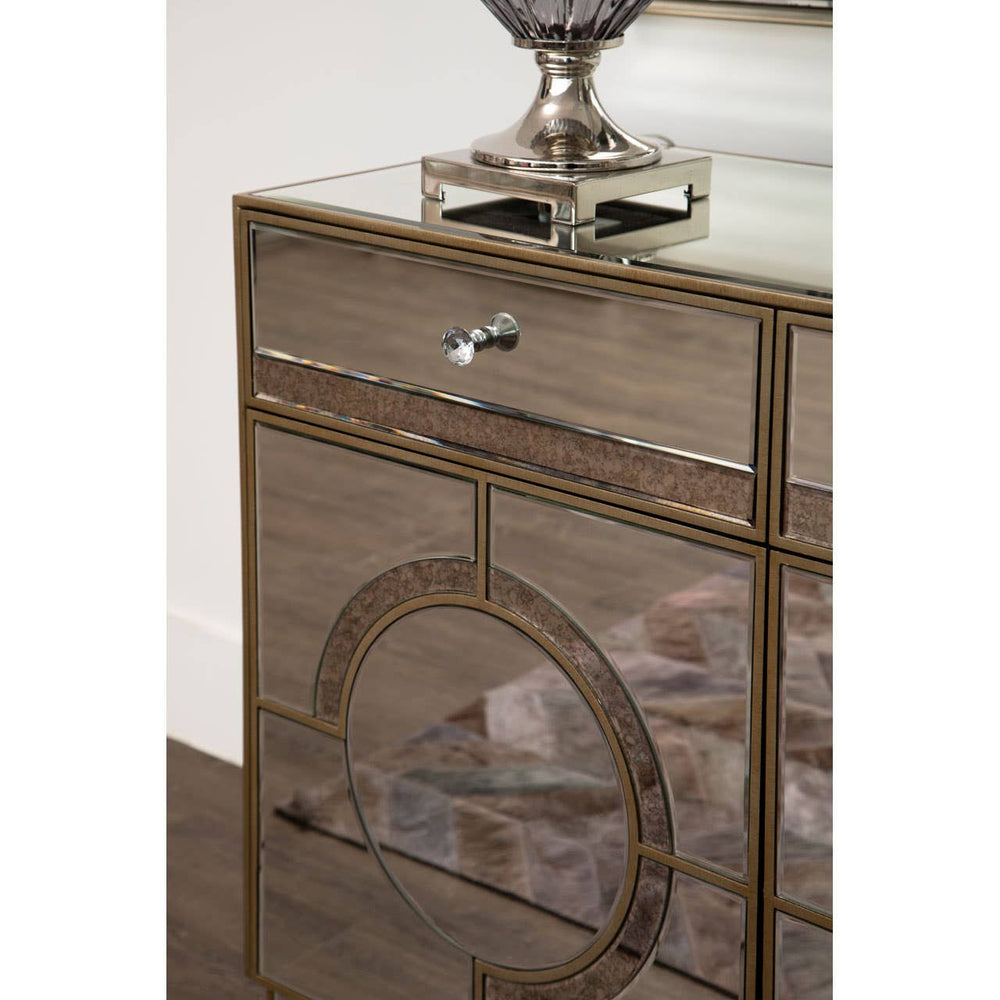 Champagne gold and mirrored sideboard featuring a sleek design with a single drawer and decorative circular detailing on the front, complemented by a polished surface and a stylish lamp on top. The sideboard is set against a wooden floor, enhancing its elegant appearance