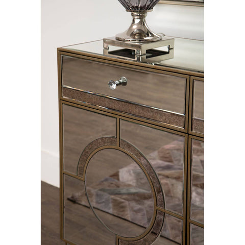 Champagne gold and mirrored sideboard featuring a sleek design with a single drawer and decorative circular detailing on the front, complemented by a polished surface and a stylish lamp on top. The sideboard is set against a wooden floor, enhancing its elegant appearance