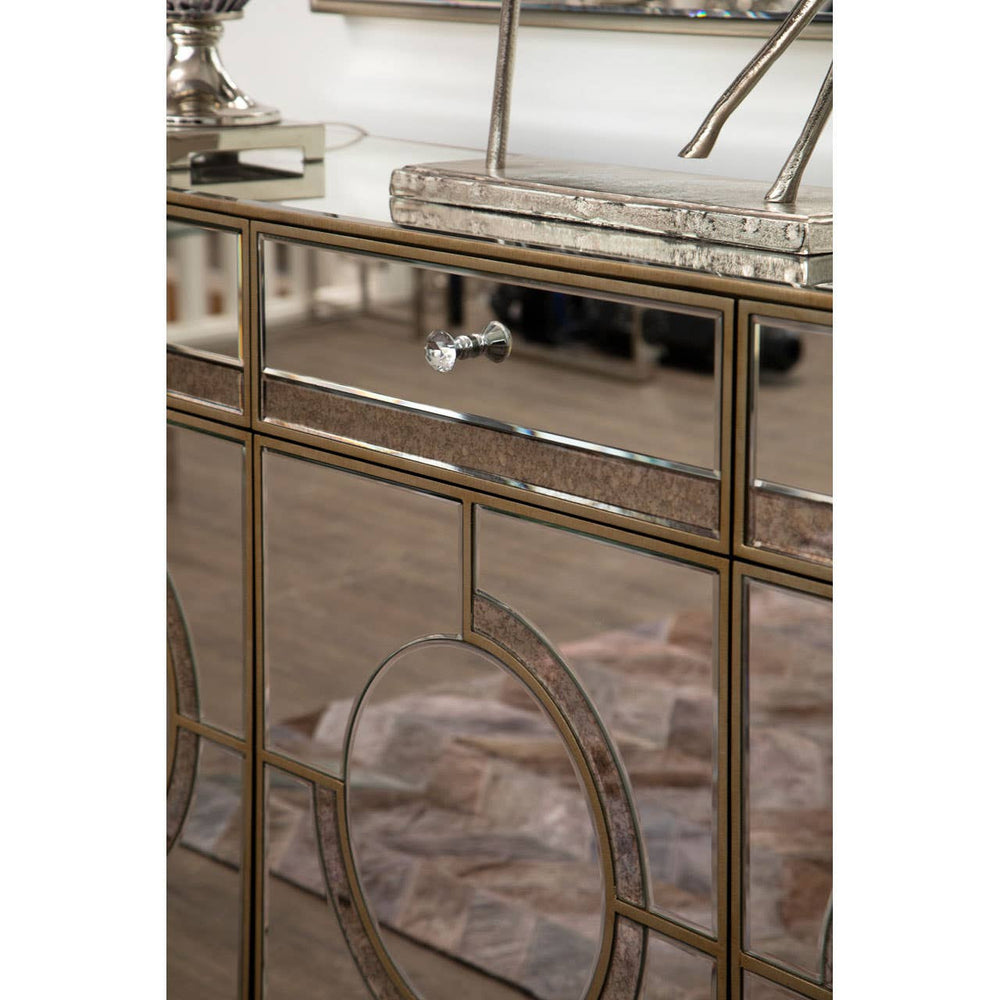 Champagne gold and mirrored sideboard featuring a geometric design, with a sleek surface and elegant detailing. The sideboard is positioned in a stylish interior setting, showcasing a soft, neutral-colored rug beneath it