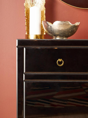 A close-up view of the Kensington Townhouse Black Cabinet featuring a sleek black finish, two drawers with elegant gold handles, and a decorative silver bowl placed on top against a warm, reddish wall