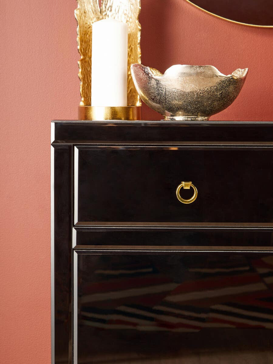 A close-up view of the Kensington Townhouse Black Cabinet featuring a sleek black finish, two drawers with elegant gold handles, and a decorative silver bowl placed on top against a warm, reddish wall