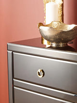 Black cabinet with a smooth surface, featuring a round gold handle on a drawer, set against a warm pink wall. A decorative bowl and two candles are placed on top of the cabinet, adding a touch of elegance to the scene