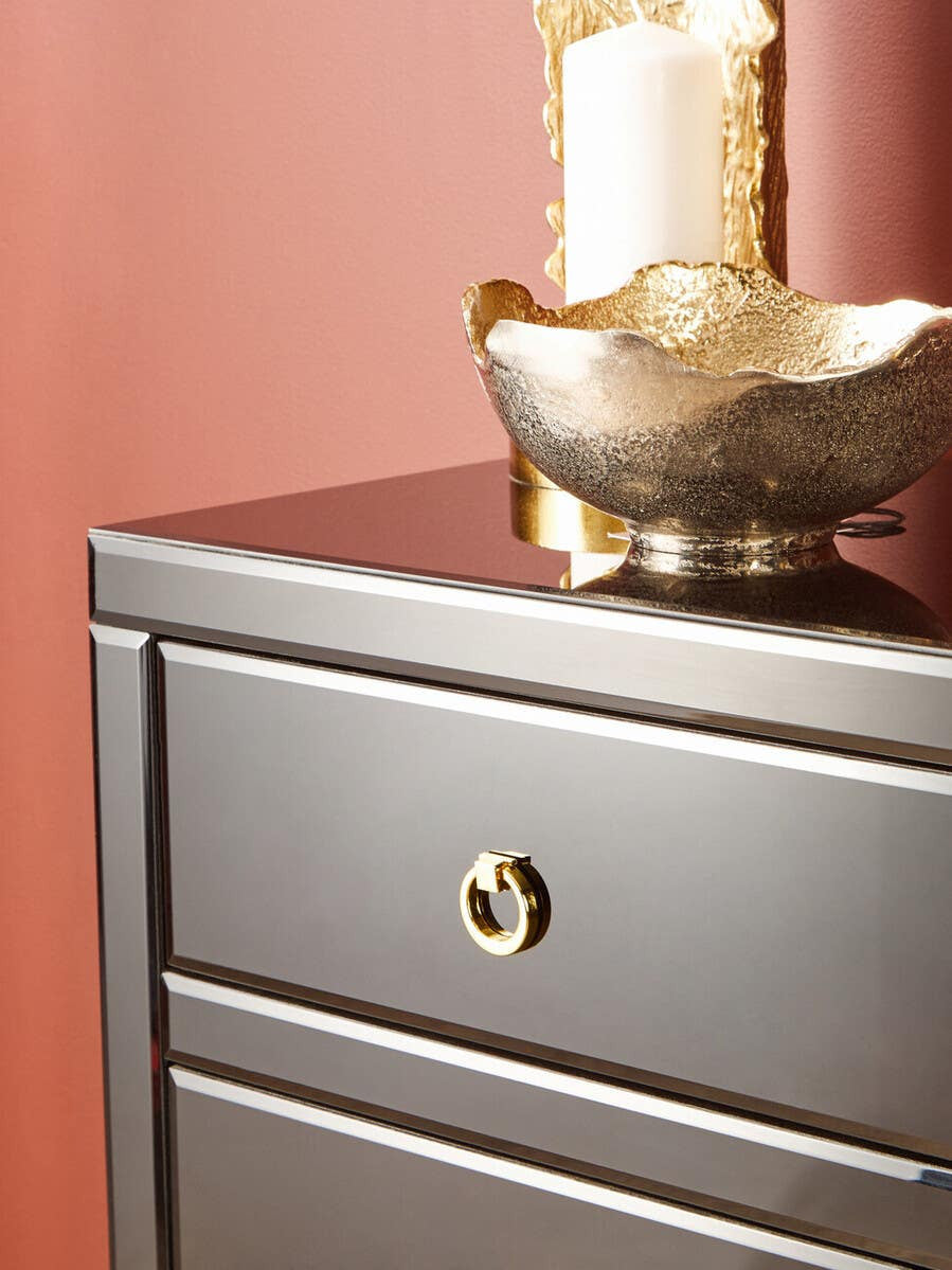 Black cabinet with a smooth surface, featuring a round gold handle on a drawer, set against a warm pink wall. A decorative bowl and two candles are placed on top of the cabinet, adding a touch of elegance to the scene