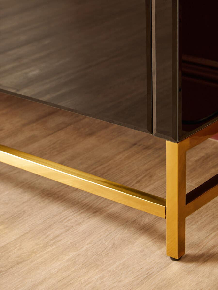 Black cabinet with a sleek design featuring a gold metal base, set against a light wooden floor. The cabinet has a smooth finish and a modern aesthetic