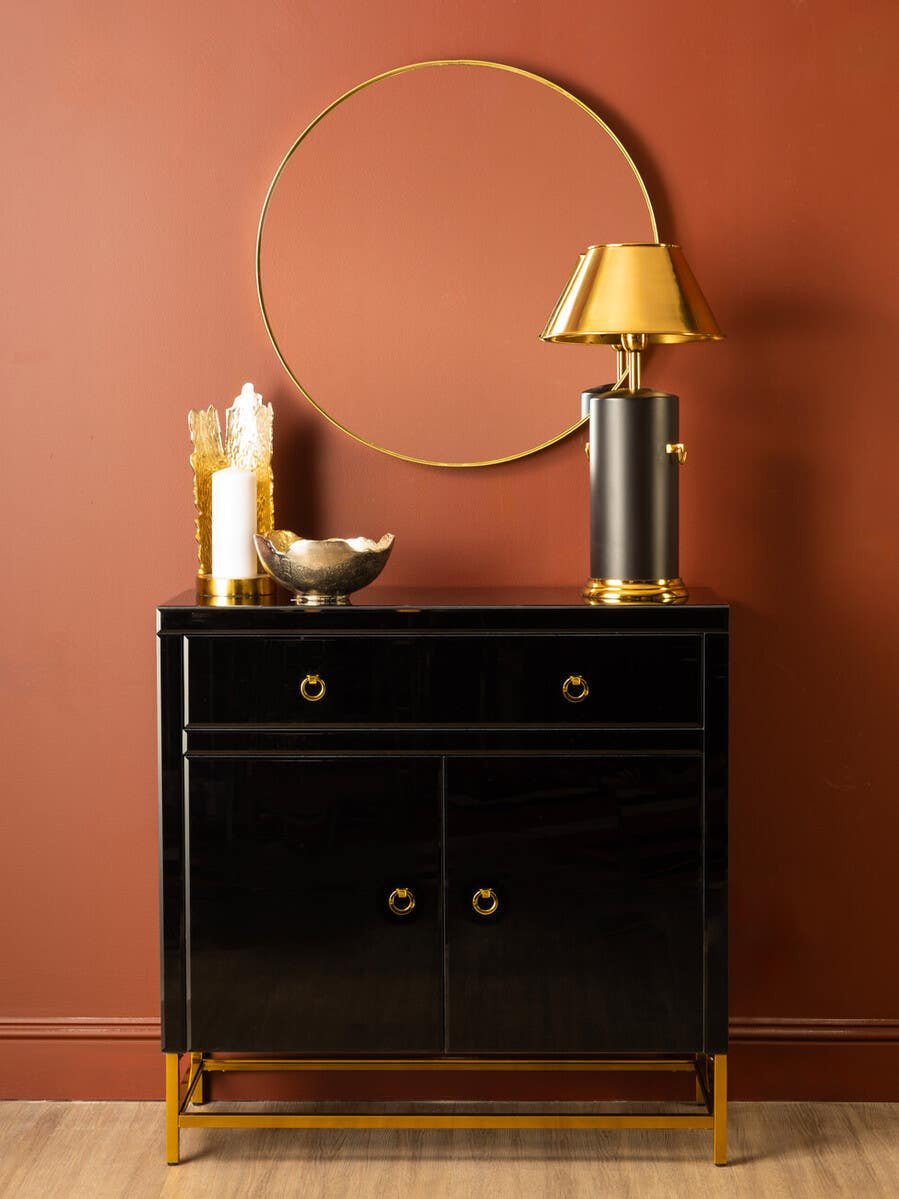 A sleek black cabinet with two drawers and two doors, featuring gold handles, sits against a warm, reddish-brown wall. On top of the cabinet, there is a round gold mirror, a small lamp with a yellow shade, a decorative bowl, and a tall black and gold bookend. The cabinet is complemented by a light wooden floor