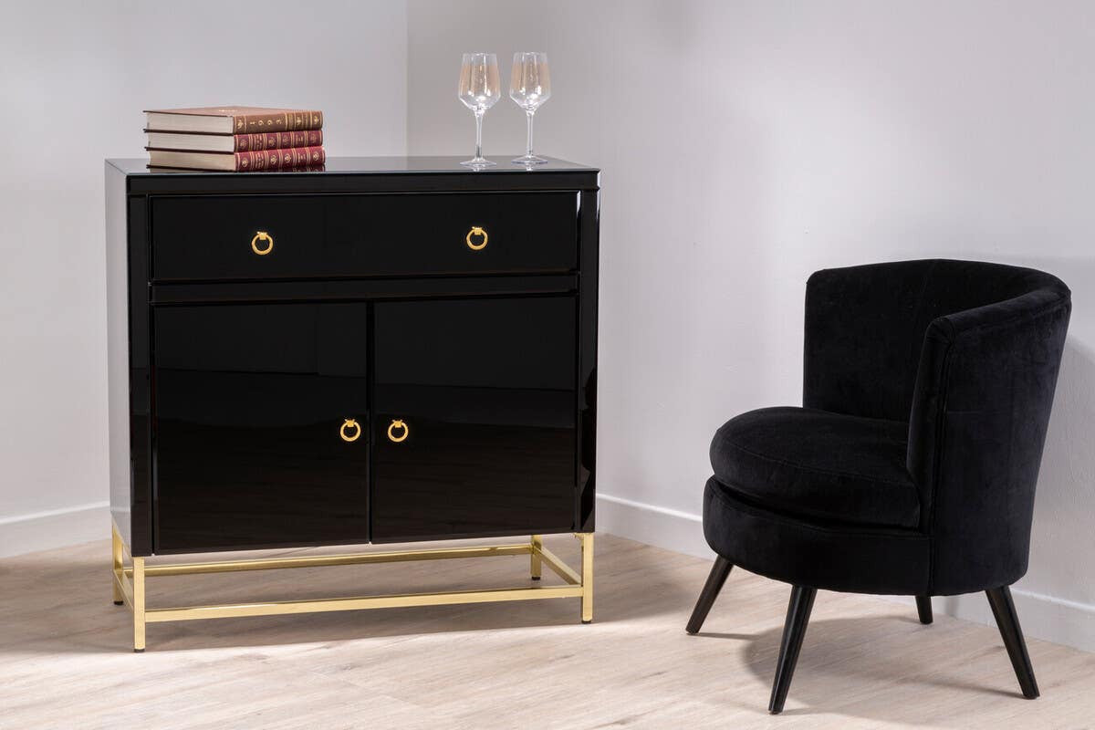 Kensington Townhouse black cabinet featuring a sleek design with three drawers and two doors, accented by gold hardware, set on a gold metal base. A plush black chair with a rounded backrest is positioned beside the cabinet, against a light-colored wall and floor. Two glasses and a decorative box are placed on top of the cabinet