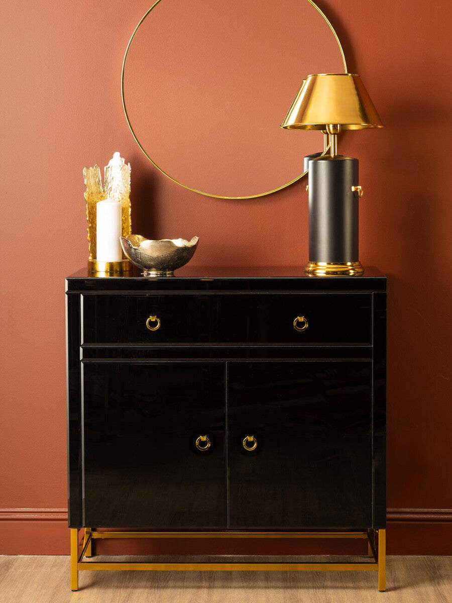 A sleek black cabinet with two drawers and two doors, featuring gold hardware and legs, positioned against a warm, terracotta wall. A round mirror hangs above the cabinet, and a stylish lamp and decorative bowl are placed on top