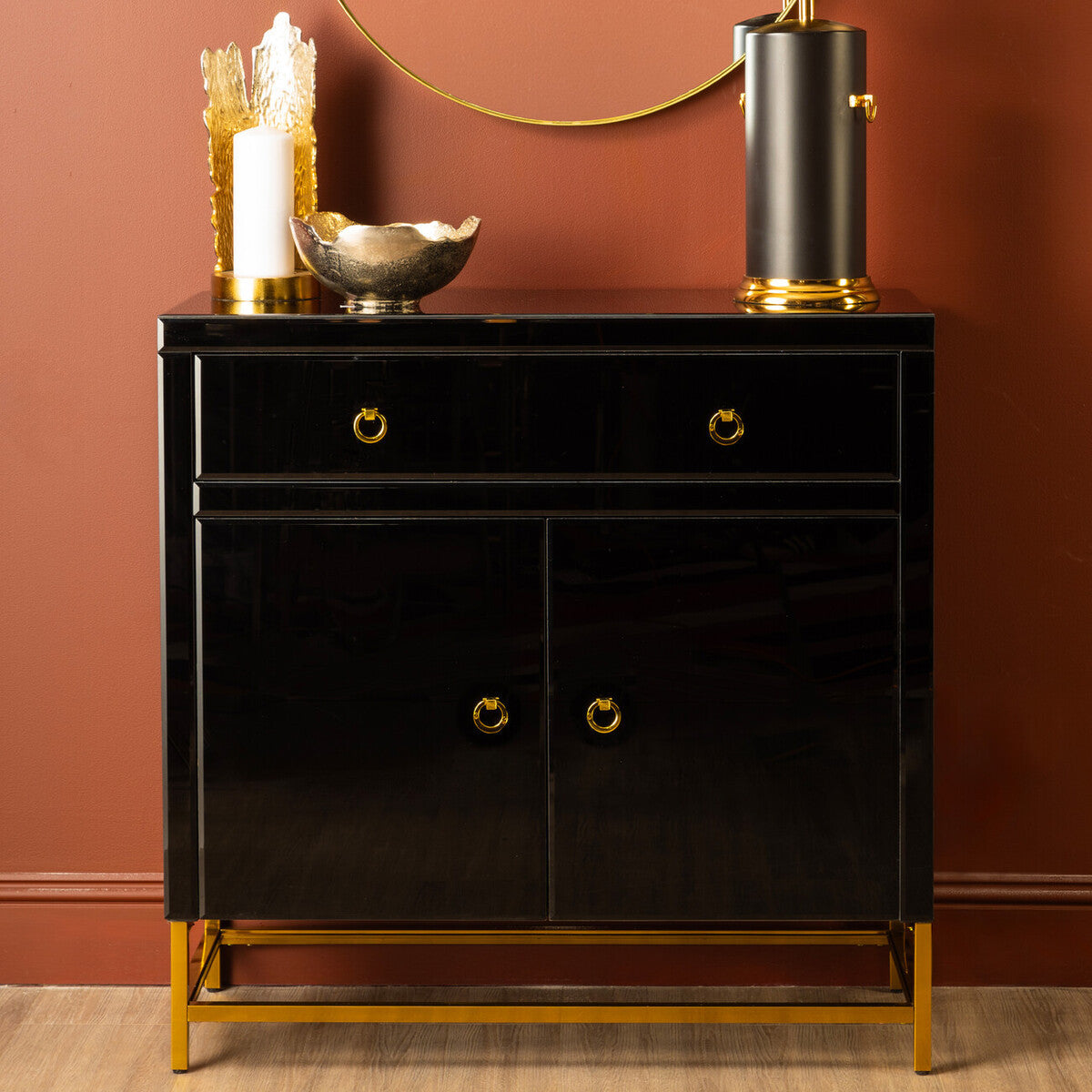 Black cabinet with two drawers and two doors, featuring gold hardware and legs. The cabinet is styled with decorative items, including a metallic vase, a bowl, and a tall, cylindrical object, set against a warm, reddish-brown wall