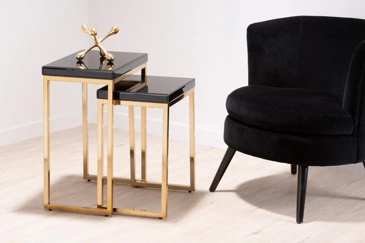 Set of two black glass nesting tables with gold metal frames beside a black velvet chair