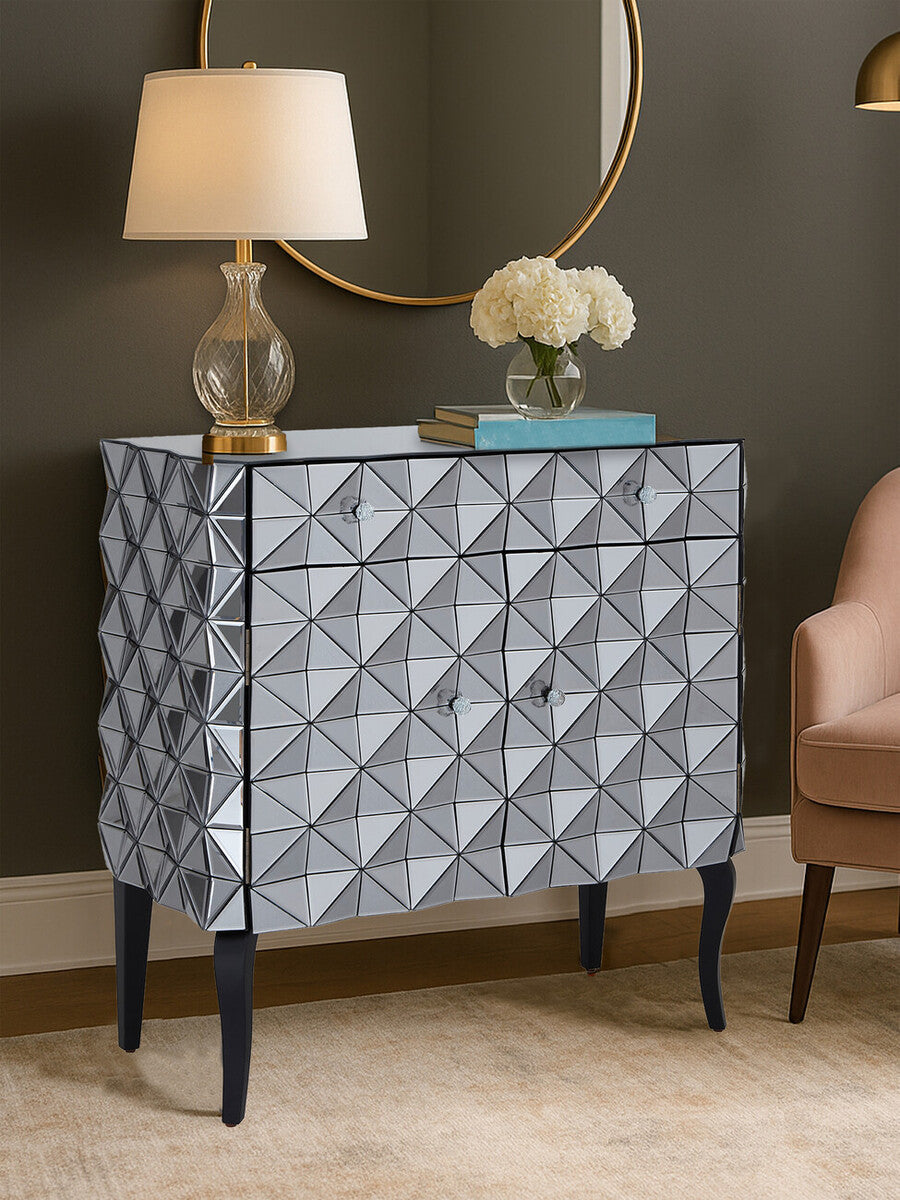 A stylish sideboard with a silver finish featuring a diamond-patterned design on the front, two doors, and sleek black legs. A lamp and a vase with flowers are placed on top, against a neutral wall and a round mirror in the background. A light-colored chair is visible to the right