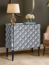 A stylish sideboard with a silver finish featuring a diamond-patterned design on the front, two doors, and sleek black legs. A lamp and a vase with flowers are placed on top, against a neutral wall and a round mirror in the background. A light-colored chair is visible to the right