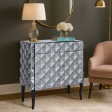 A stylish sideboard with a silver finish featuring a diamond-patterned design, two doors, and sleek black legs. The sideboard is placed against a dark wall, accompanied by a lamp and a small vase of flowers on top, with a light-colored armchair nearby