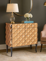 A stylish two-door cabinet with a copper finish, featuring a diamond pattern on the front, black legs, and decorative hardware. A lamp and a vase with flowers sit on top, alongside a stack of books, against a dark wall. A light-colored chair is partially visible on the right