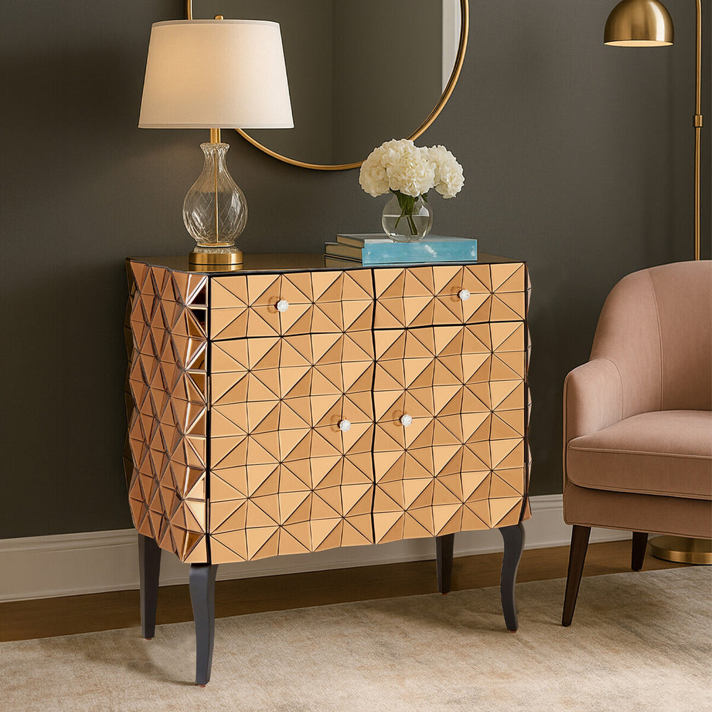 Soho 2 Door Copper Finish Cabinet featuring a textured, diamond-patterned front, two doors with sleek handles, and elegant black legs. The cabinet is positioned next to a soft pink armchair and a round mirror, with a lamp and decorative flowers on top, set against a dark wall