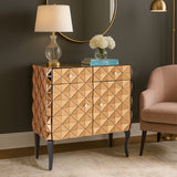 Soho 2 Door Copper Finish Cabinet featuring a textured, diamond-patterned front, two doors with sleek handles, and elegant black legs. The cabinet is positioned next to a soft pink armchair and a round mirror, with a lamp and decorative flowers on top, set against a dark wall