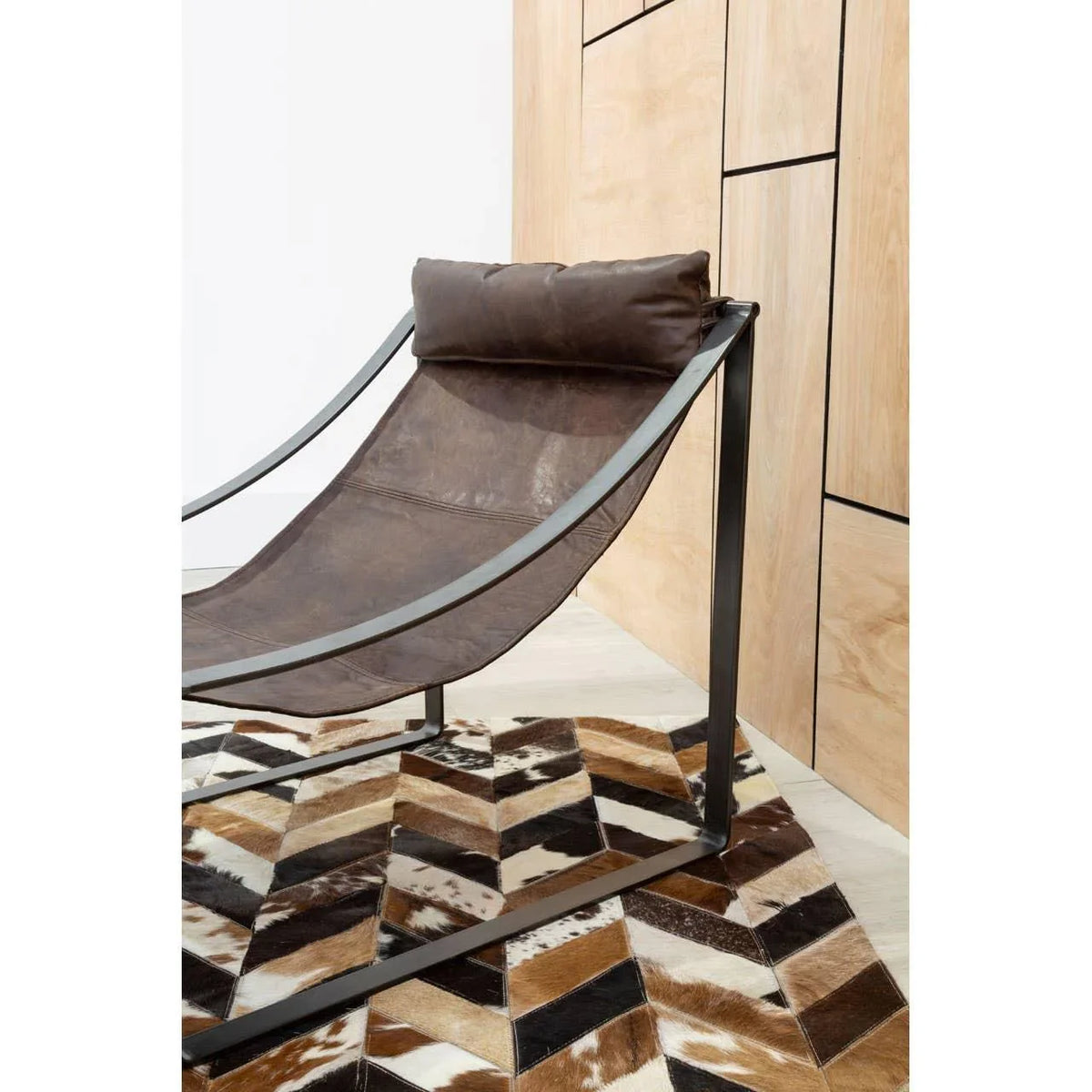 Brown leather lounge chair with metal frame on geometric cowhide rug by wood panel wall
