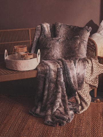 A cozy woven rope sofa with three seats, featuring plush cushions and a textured fabric, is positioned against a warm-toned wall. A soft, patterned throw blanket drapes over one arm of the sofa, while a round basket filled with decorative items sits nearby on a wooden surface. The flooring is a rich wood finish, enhancing the inviting atmosphere of the space