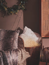 A stylish three-seater sofa featuring woven rope detailing, adorned with plush cushions in various textures. The setting includes a warm, inviting atmosphere with soft lighting and decorative elements in the background, such as a wooden cabinet and greenery