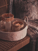 A cozy arrangement featuring a woven rope sofa with a soft, textured throw draped over it. A round tray sits on a wooden surface, holding two decorative glass containers with natural fiber accents, surrounded by a plush, dark fur blanket. The warm color palette creates an inviting atmosphere