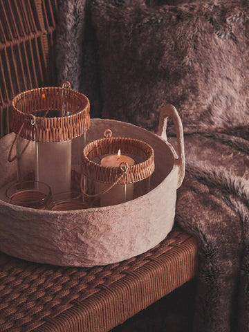 A cozy arrangement featuring a woven rope sofa with a textured fabric throw draped over it. A round tray holds two glass candle holders with natural wooden accents, set on a wooden surface. The background includes soft, plush textiles, creating a warm and inviting atmosphere