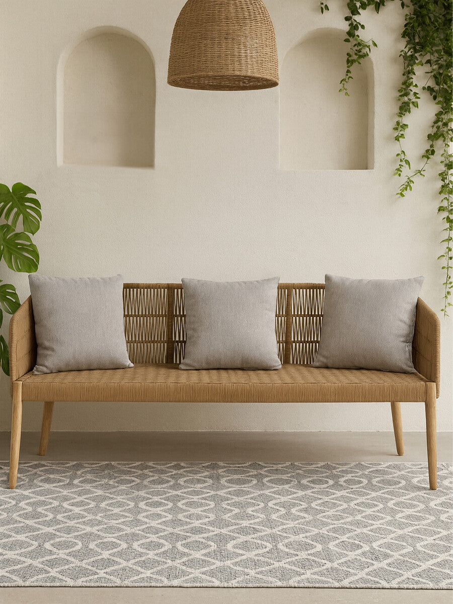 A three-seater sofa featuring a woven rope backrest and wooden frame, complemented by three light gray cushions. The sofa is positioned in a bright room with a patterned rug, a hanging light fixture, and green plants in the background