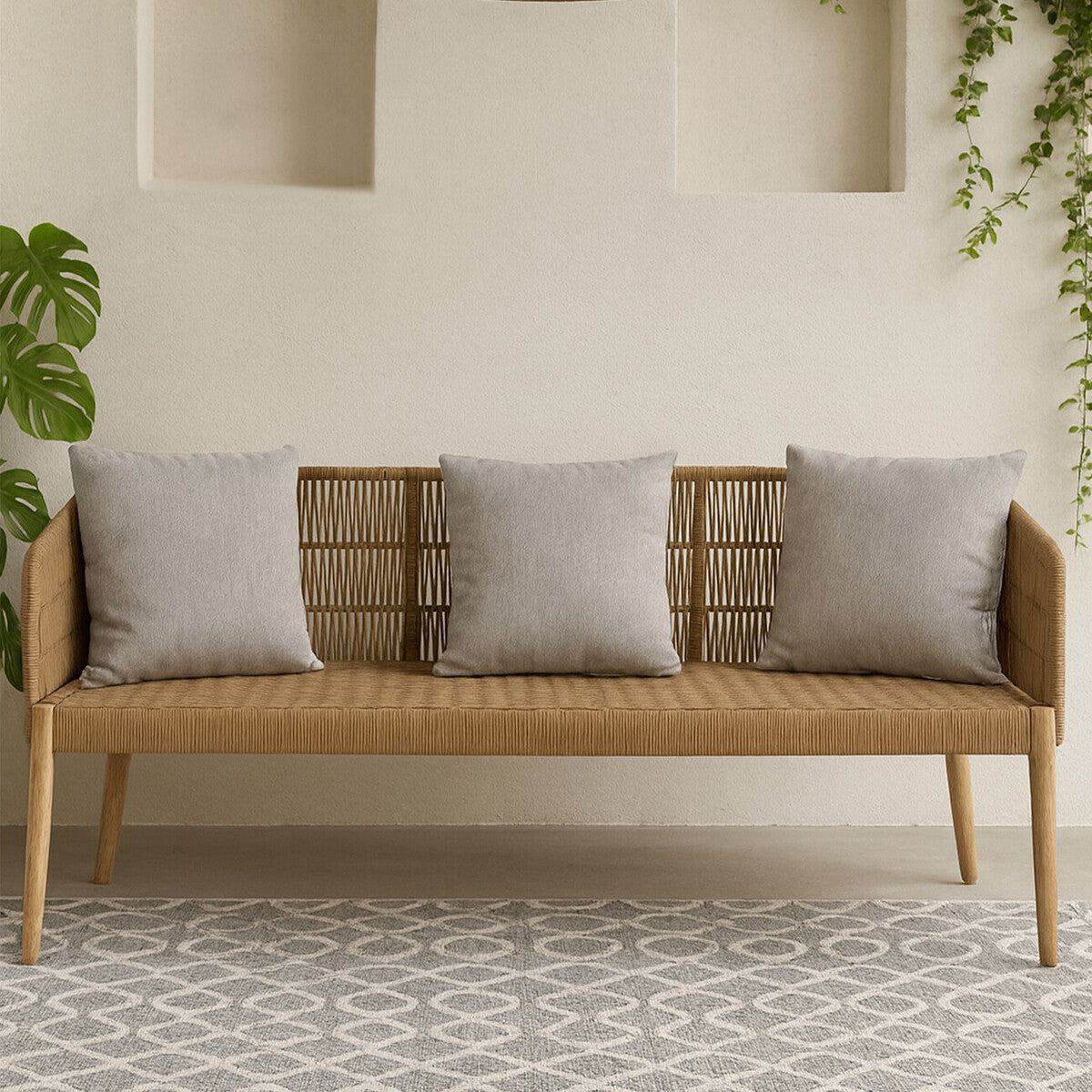 Three-seater sofa featuring a woven rope backrest and wooden frame, complemented by four light gray cushions. The sofa is positioned against a neutral wall with decorative niches, and a patterned rug is visible beneath it. Green foliage is present in the background, adding a touch of nature to the setting
