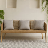 Three-seater sofa featuring a woven rope backrest and wooden frame, complemented by four light gray cushions. The sofa is positioned against a neutral wall with decorative niches, and a patterned rug is visible beneath it. Green foliage is present in the background, adding a touch of nature to the setting