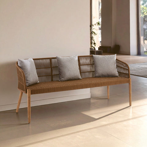 A three-seater sofa featuring a woven rope design with a wooden frame and legs. The sofa is adorned with three light gray cushions, positioned against a neutral wall in a bright, airy room with natural light streaming in. The floor is a light-colored material, enhancing the modern aesthetic of the space