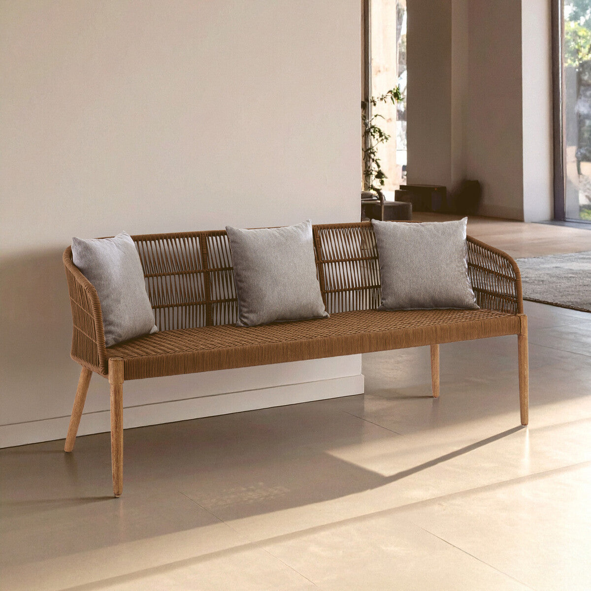 A three-seater sofa featuring a woven rope design with a wooden frame and legs. The sofa is adorned with three light gray cushions, positioned against a neutral wall in a bright, airy room with natural light streaming in. The floor is a light-colored material, enhancing the modern aesthetic of the space