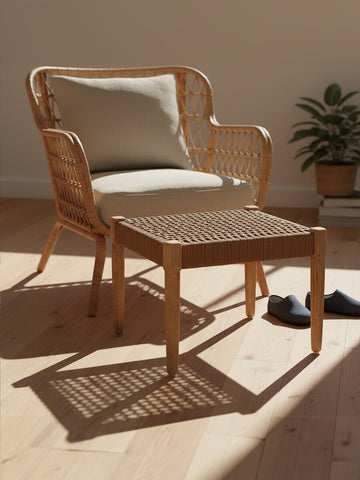 Woven rope footstool with a square shape, featuring a natural wood frame and a textured surface, placed next to a light-colored armchair with a cushion. The setting includes a warm wooden floor and a potted plant in the background, illuminated by soft natural light