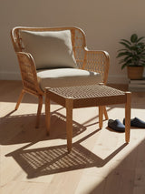 Woven rope footstool with a square shape, featuring a natural wood frame and a textured surface, placed next to a light-colored armchair with a cushion. The setting includes a warm wooden floor and a potted plant in the background, illuminated by soft natural light