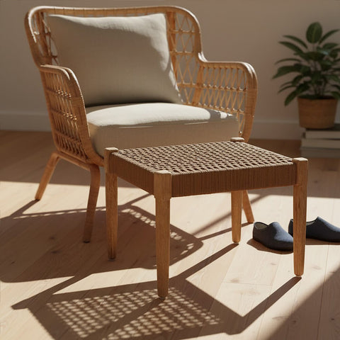 Woven rope footstool in a natural finish, placed next to a light-colored armchair with a beige cushion. The footstool features a square design with a textured surface, and the scene is illuminated by soft, natural light, highlighting the wooden floor and a small potted plant in the background. A pair of dark slippers is positioned beside the footstool
