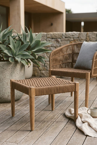 Woven rope footstool with a natural wood frame, placed on a wooden deck beside a chair and a large potted plant. The footstool features a minimalist design with a slatted top and four sturdy legs. A cozy blanket is draped on the floor nearby