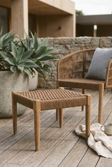 Woven rope footstool with a natural wood frame, placed on a wooden deck beside a chair and a large potted plant. The footstool features a minimalist design with a slatted top and four sturdy legs. A cozy blanket is draped on the floor nearby