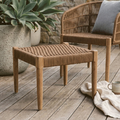 Woven rope footstool with a square top and four sturdy legs, featuring a natural brown color and textured surface, placed on a wooden deck beside a chair and a decorative pillow, with a plant in the background