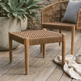 Woven rope footstool with a square top and four sturdy legs, featuring a natural brown color and textured surface, placed on a wooden deck beside a chair and a decorative pillow, with a plant in the background