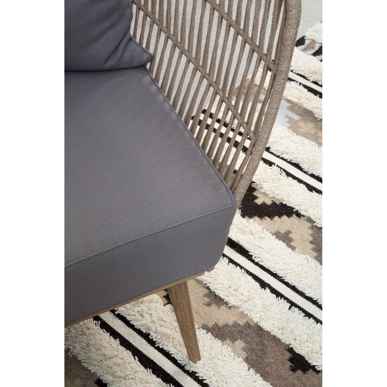 Woven rope two-seater sofa featuring a light brown frame and grey cushions, positioned on a patterned rug with geometric designs in neutral tones. The sofa has a curved backrest and wooden legs, showcasing a modern and stylish design