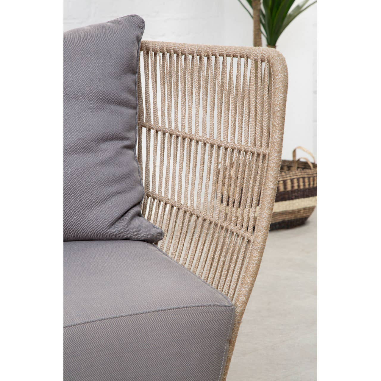 Woven rope two-seater sofa featuring a light beige frame and soft grey cushions, showcasing a modern design with a curved backrest and clean lines. The sofa is positioned in a bright, minimalistic interior setting with a hint of greenery in the background