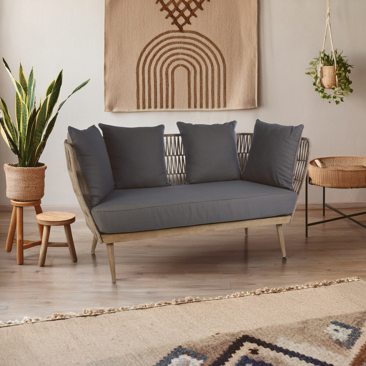 Woven rope two-seater sofa featuring a light wooden frame and four plush grey cushions, set in a cozy living space with a decorative wall hanging, a potted plant, and a patterned rug
