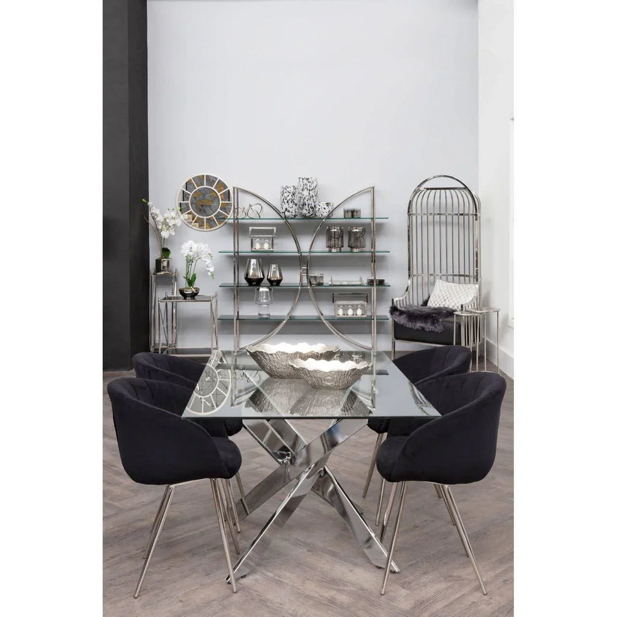 Glass dining table with chrome base and black chairs in modern room with shelving and decor