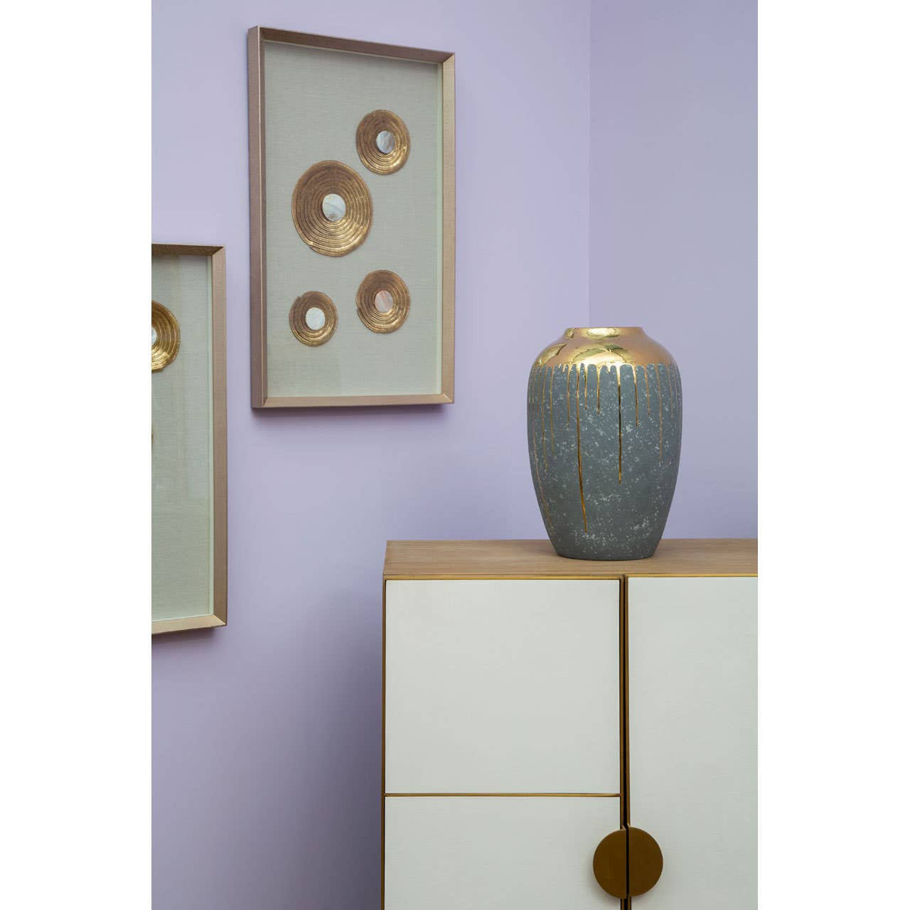 A stylish cabinet featuring a combination of oak wood and brushed gold accents, positioned against a light purple wall. The cabinet has a smooth white finish with a circular gold handle, and a decorative gray vase with a gold top sits on its surface. Two framed wall art pieces with circular designs in gold and white are displayed above the cabinet